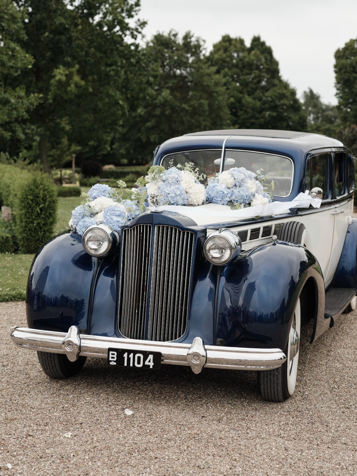 A dark blue vintage car decorated with white and blue hydrangea flowers on the hood.