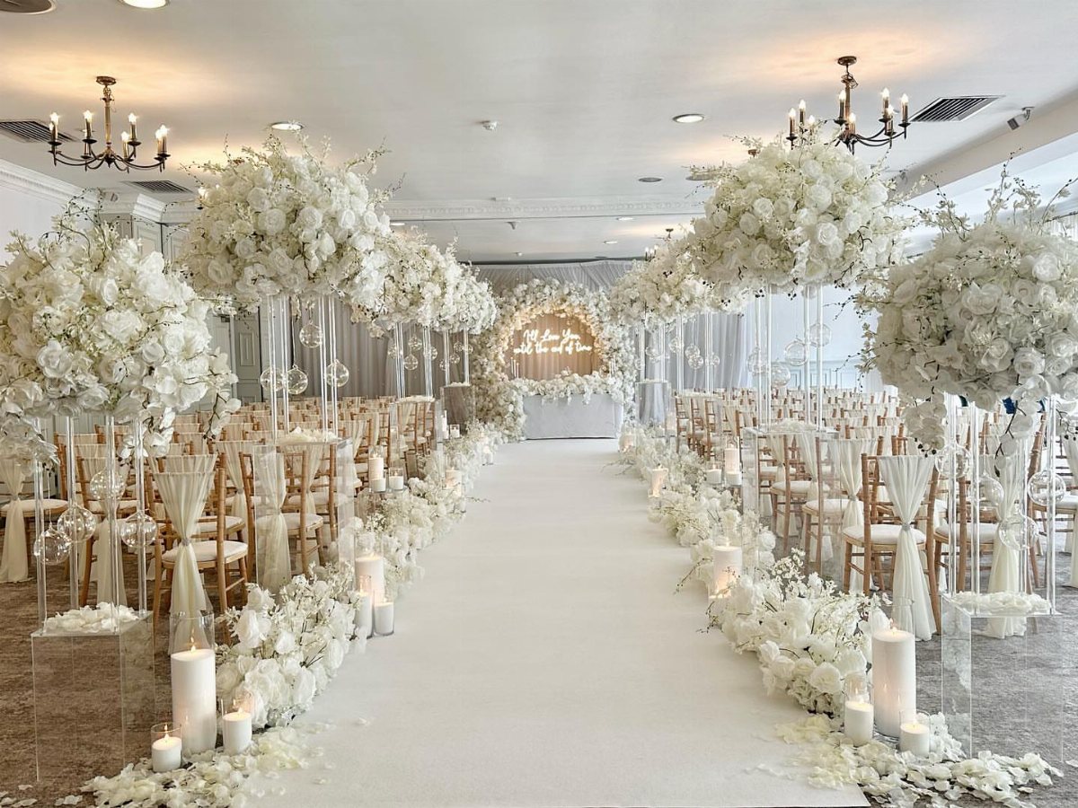 A white wedding aisle lined with large floral arrangements and candles leading to an arch.