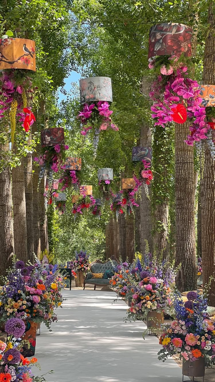 Hanging floral lamps and large colorful flower arrangements line a tree-shaded walkway.
