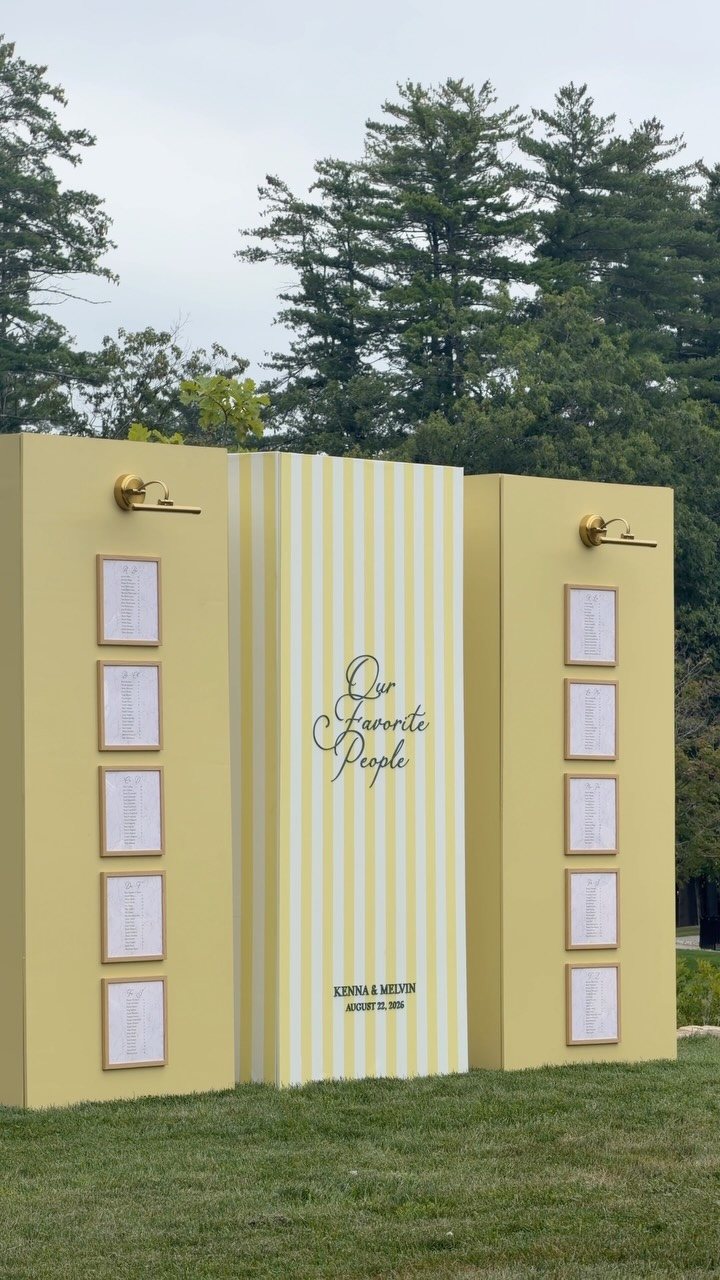 A large yellow three-panel wedding sign with cursive text and framed guest lists on grass.