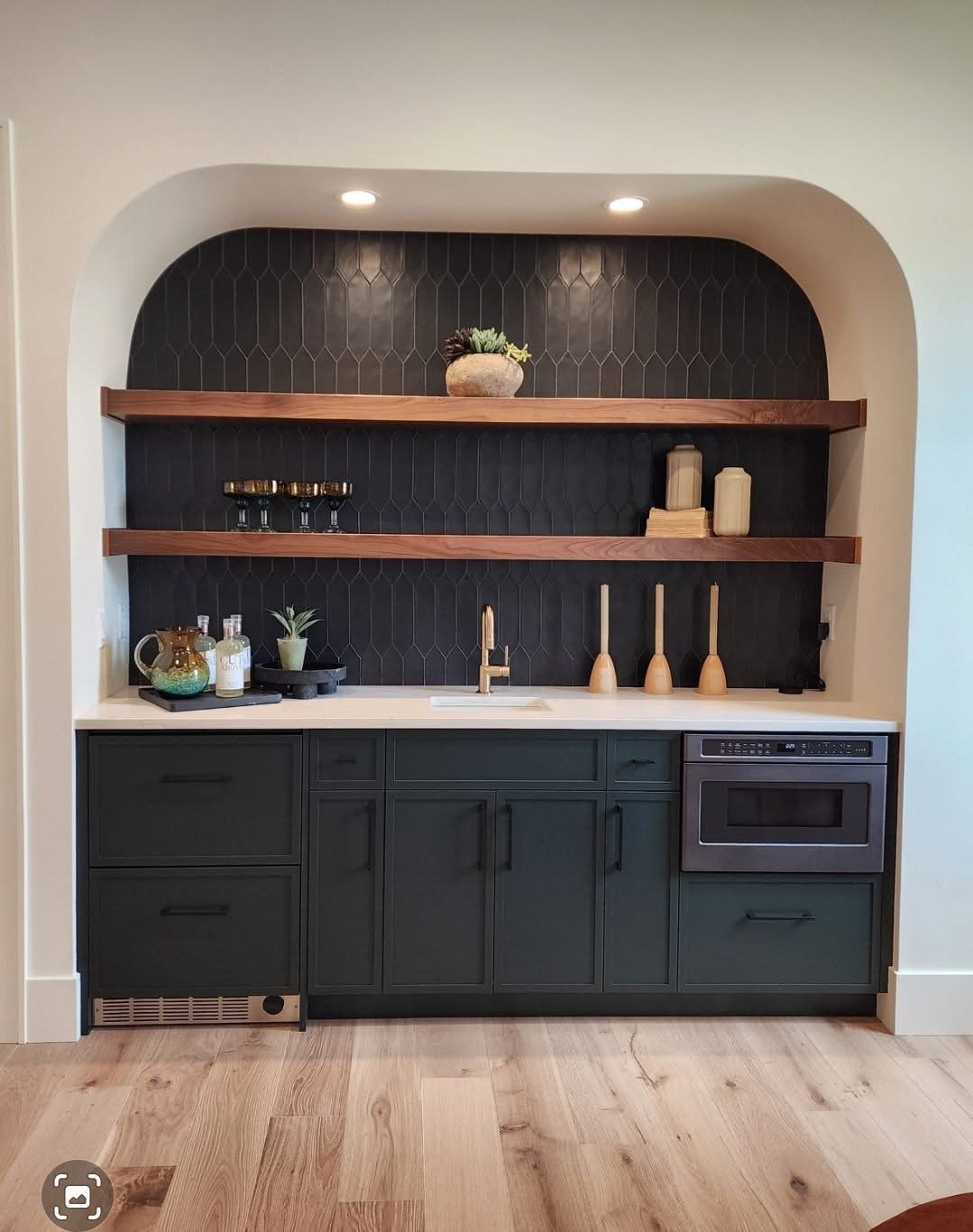 17 Basement Kitchenette Ideas That Maximize Every Square Foot 15 Dark green cabinetry and wooden shelves set within a white arched alcove with a dark tiled backsplash.