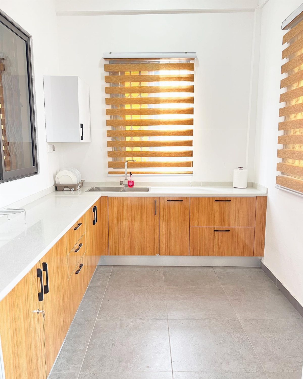 17 Basement Kitchenette Ideas That Maximize Every Square Foot 1 L-shaped kitchenette with light wood cabinets, white countertops, and a stainless steel sink under a window.