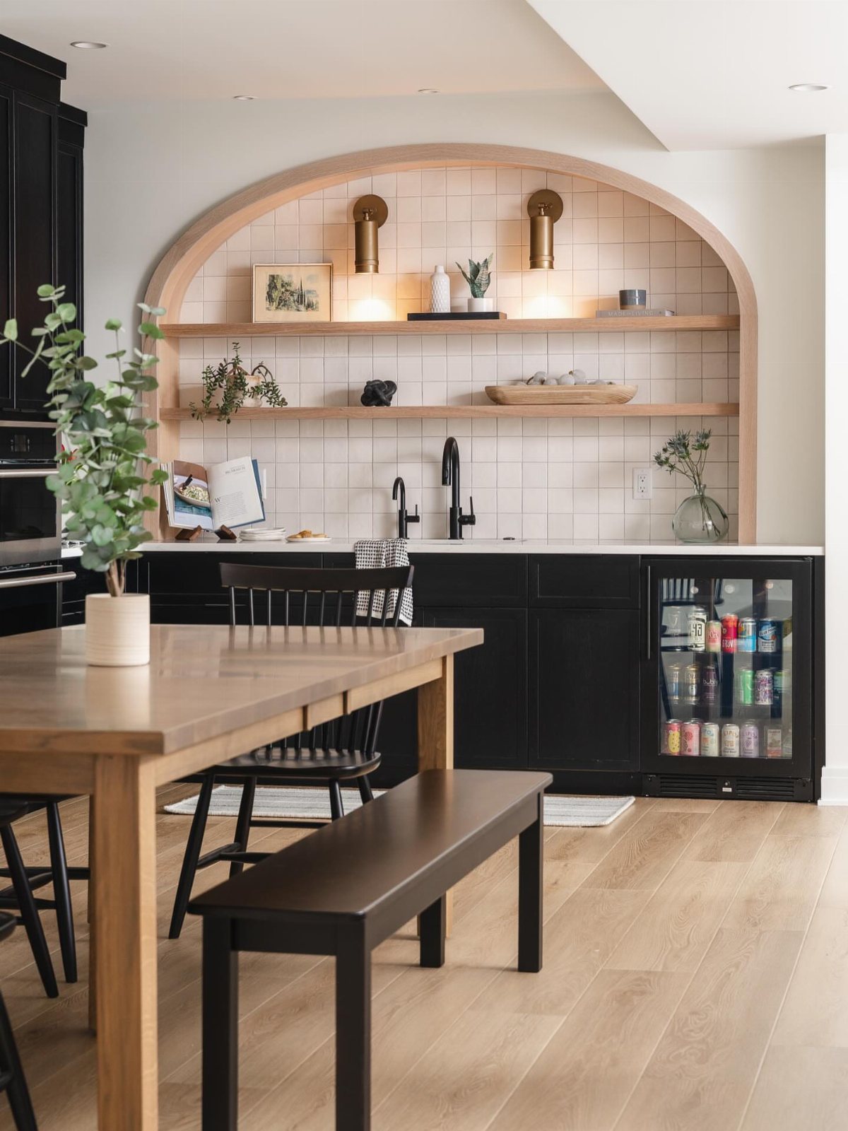 17 Basement Kitchenette Ideas That Maximize Every Square Foot 6 Modern kitchenette with black cabinets, white tile backsplash, and light wood shelving under an arch.