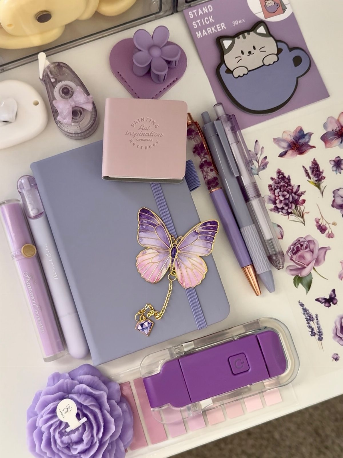 Purple pens, a lavender notebook, and decorative butterfly charms arranged on a white desk surface.