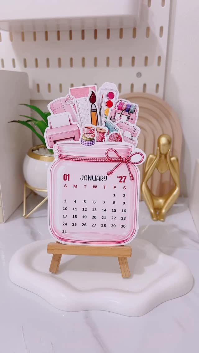 Pink makeup jar shaped calendar standing on a small wooden easel on a white tray.