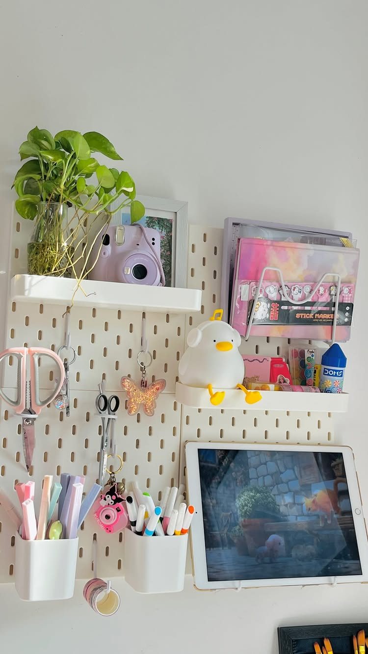 11 Desk Organization Ideas That Tidy Your Space for 2026 8 White pegboard organizer holding a plant, pink camera, scissors, pens, and a tablet on a wall.