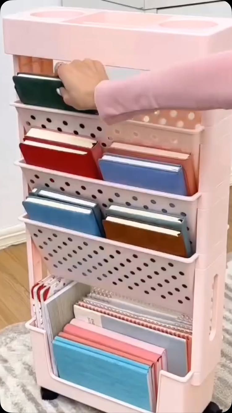 11 Desk Organization Ideas That Tidy Your Space for 2026 5 A hand organizing colorful notebooks inside a pink rolling storage cart with white bins.