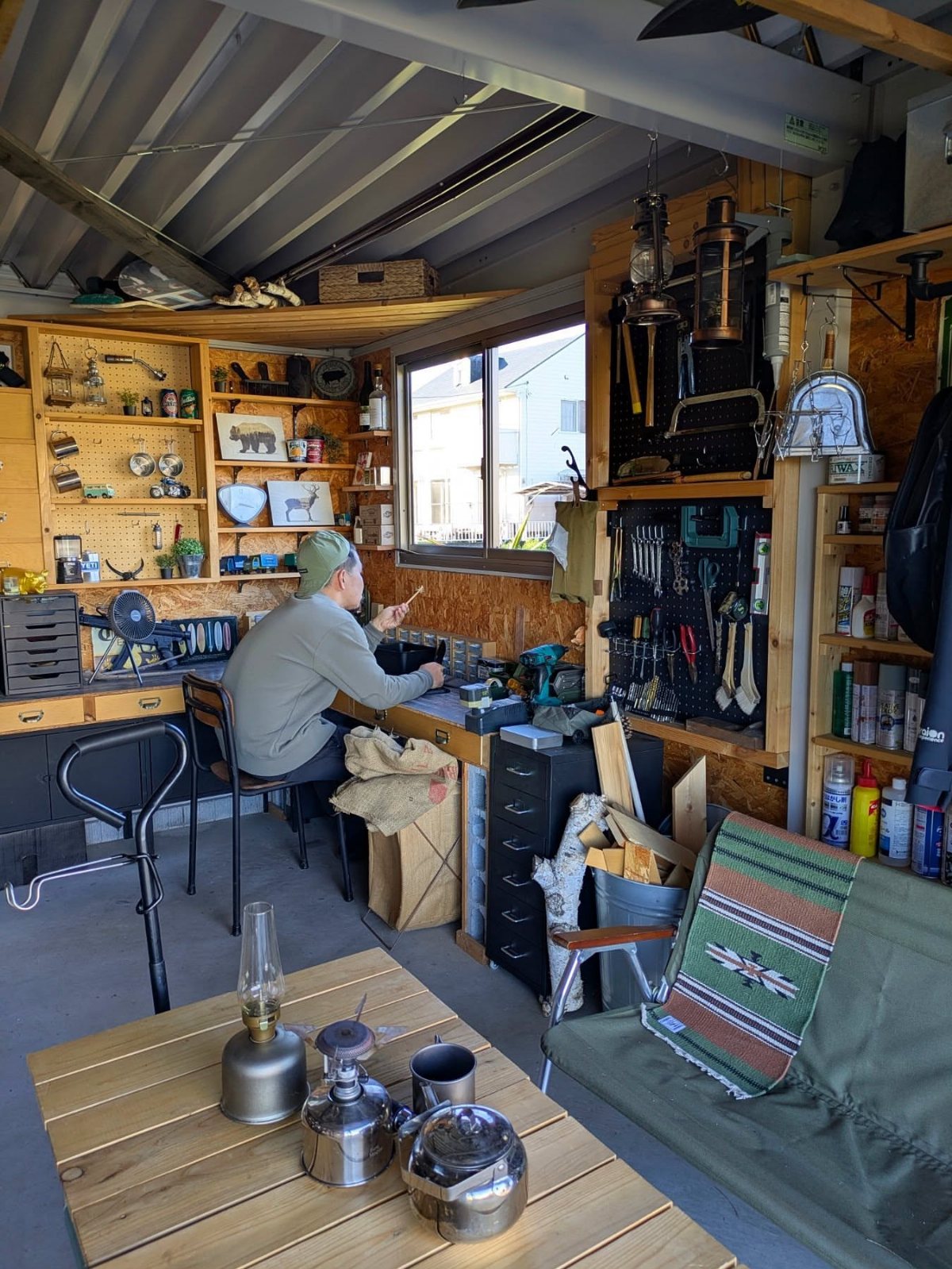 A man working at a cluttered workbench inside a wooden garage hobby workshop.