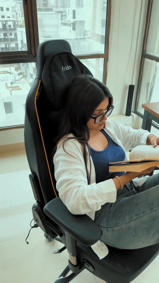 A woman sitting in a black ergonomic office chair while reading a book near a large window.