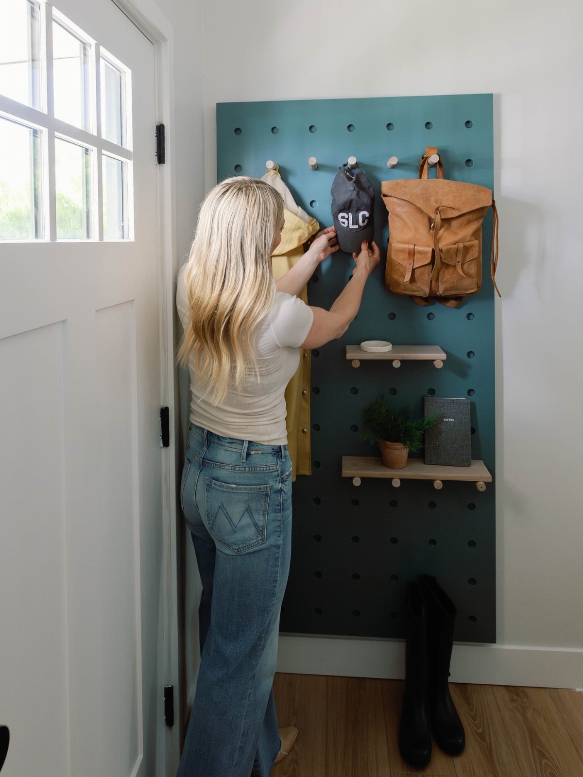 17 Mudroom Ideas for Entryways That Actually Work in 2026 13 Woman hanging a grey cap on a teal pegboard wall with wooden pegs and shelves.