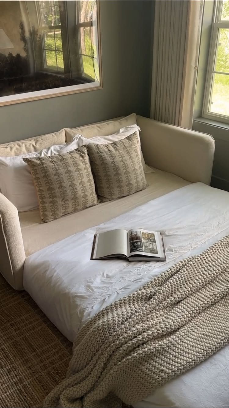 A cream convertible sofa bed styled with neutral pillows, a knit throw, and an open book.