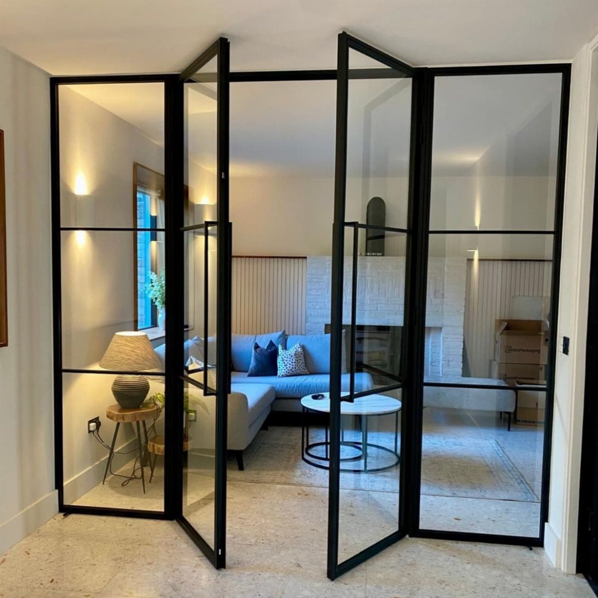 Black metal-framed glass folding doors separating a modern living room with a blue sofa.