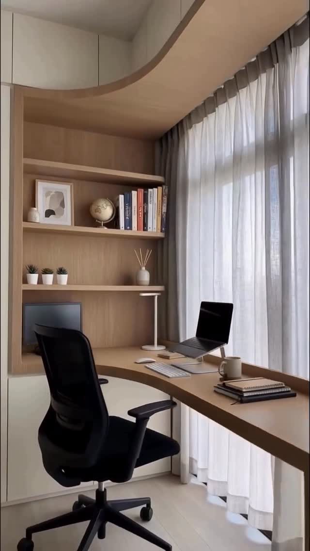 Built-in wooden desk and shelving unit positioned in front of a large window with sheer curtains.