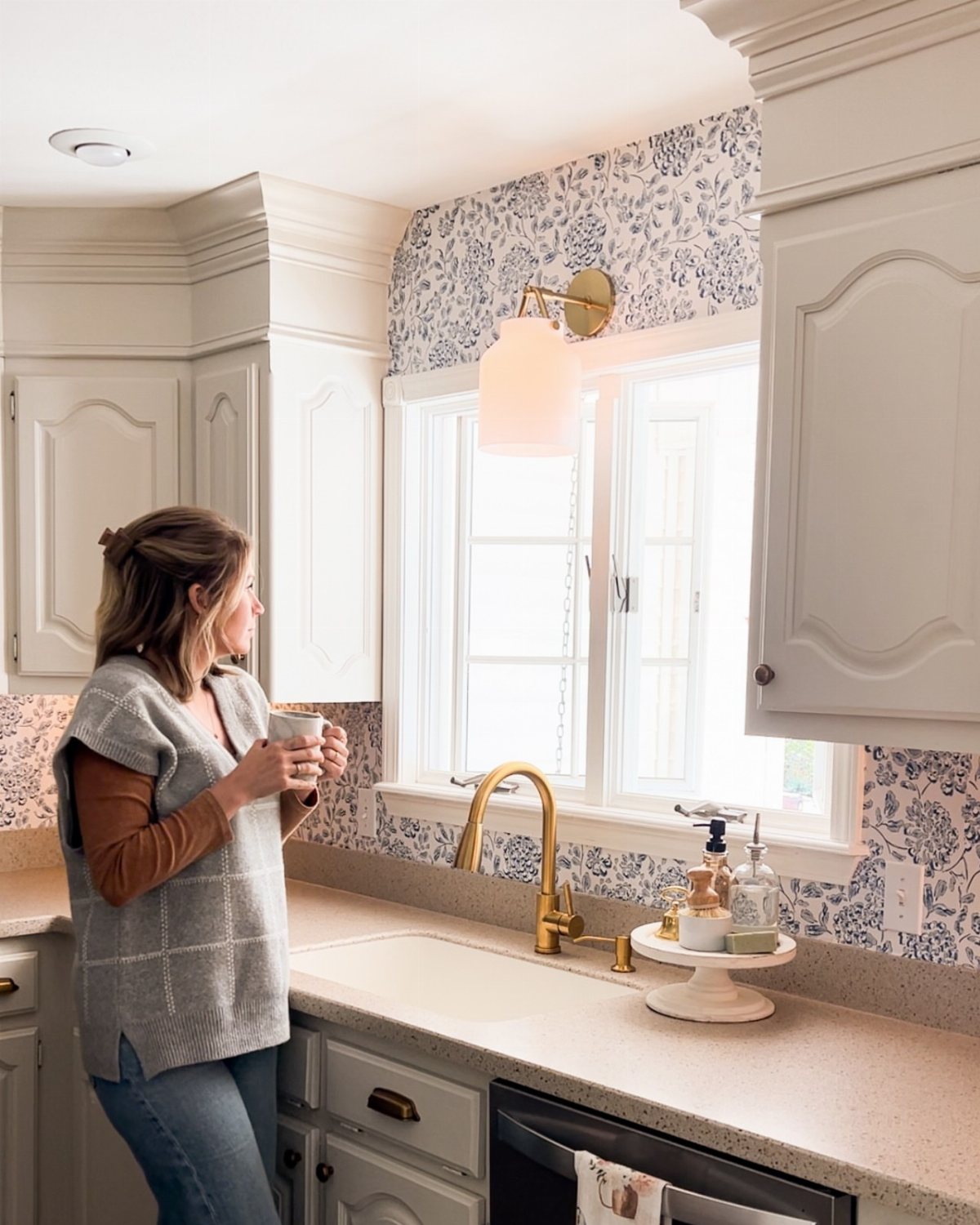 A woman in a kitchen looking out a window with floral wallpaper backsplash.