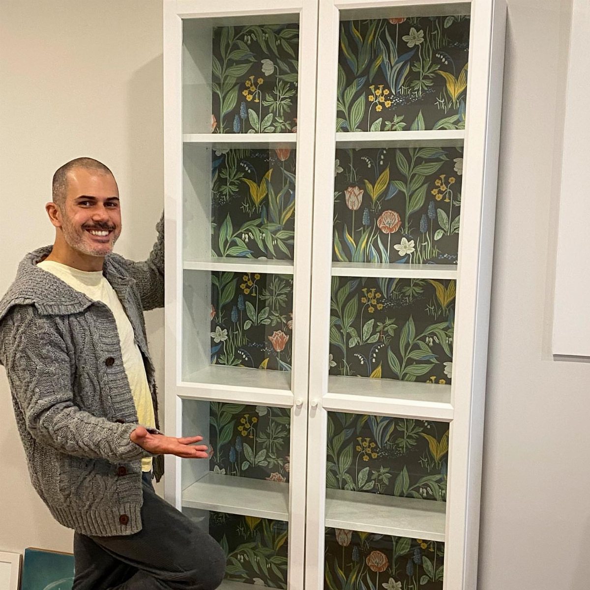 A man gestures toward a white bookcase lined with dark floral patterned wallpaper.