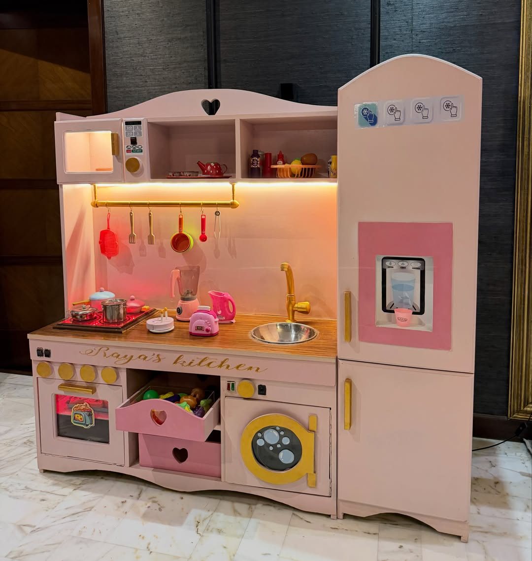 Pink wooden toy play kitchen set with miniature accessories and warm lighting.