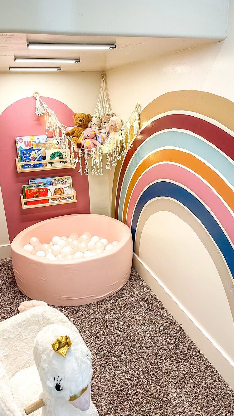 A pink ball pit sits below wall-mounted bookshelves and a macrame toy hammock.