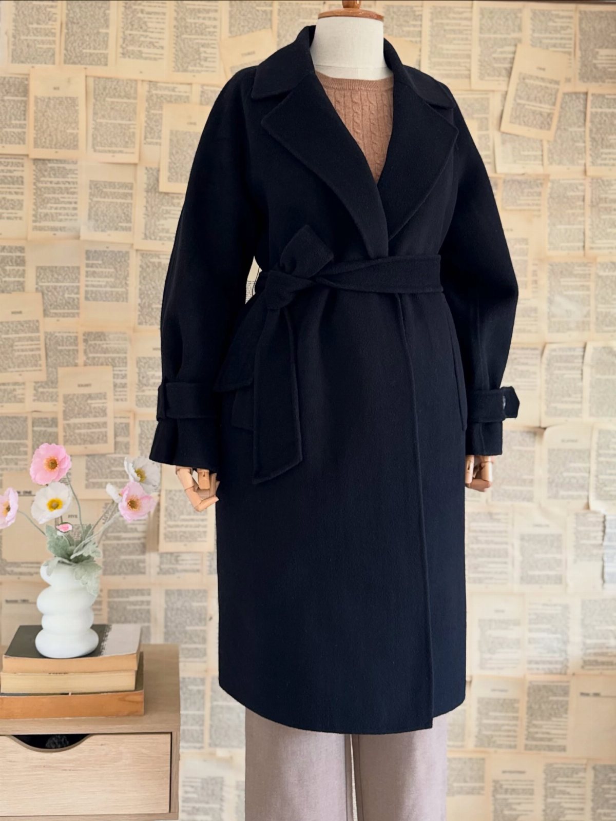 Black wrap coat with a belted waist displayed on a mannequin against a newsprint background.