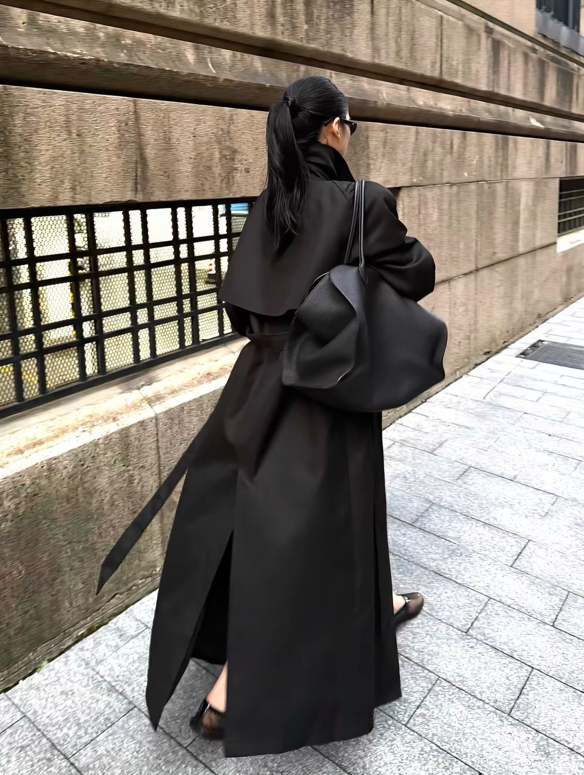 A woman in a long black wrap coat walking on a city sidewalk.
