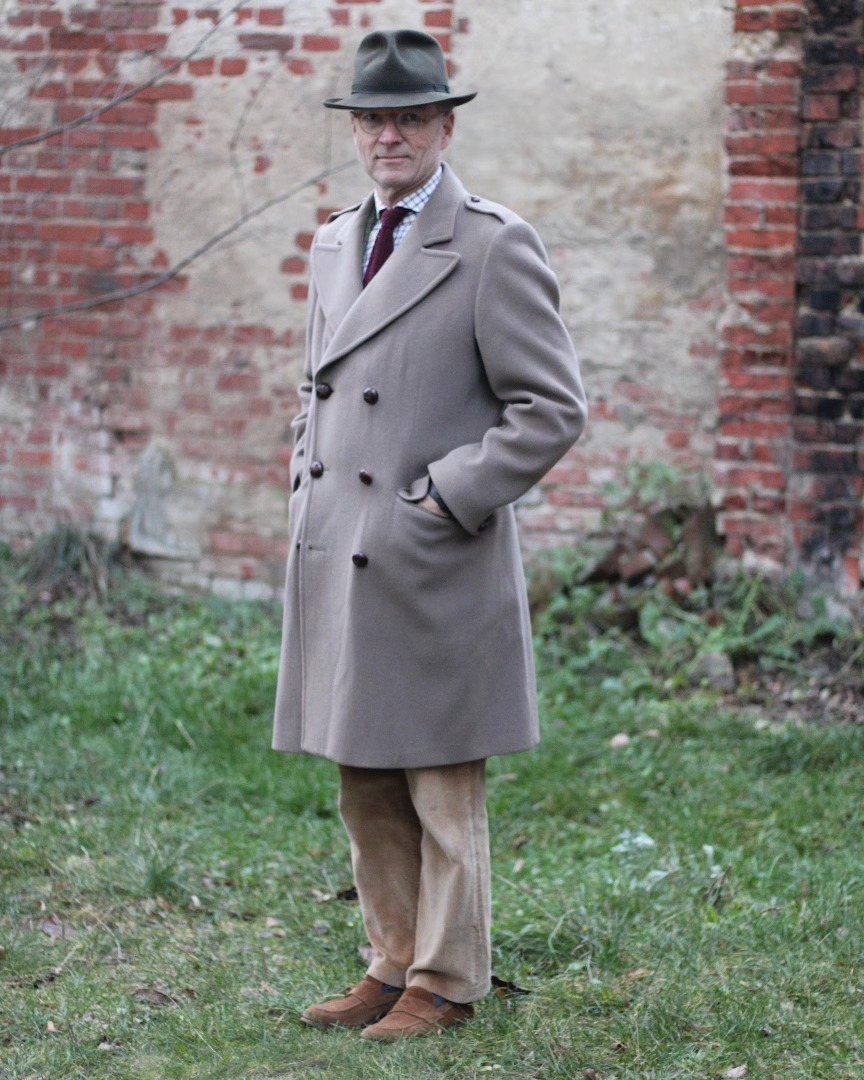 Man wearing a tan double-breasted wool overcoat and a fedora standing by a brick wall.