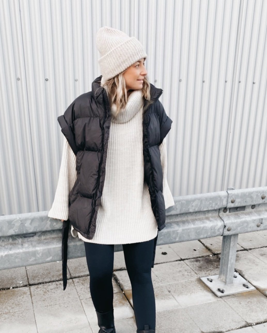 Woman wearing a cream oversized turtleneck sweater, black leggings, a black puffer vest, and a cream beanie.