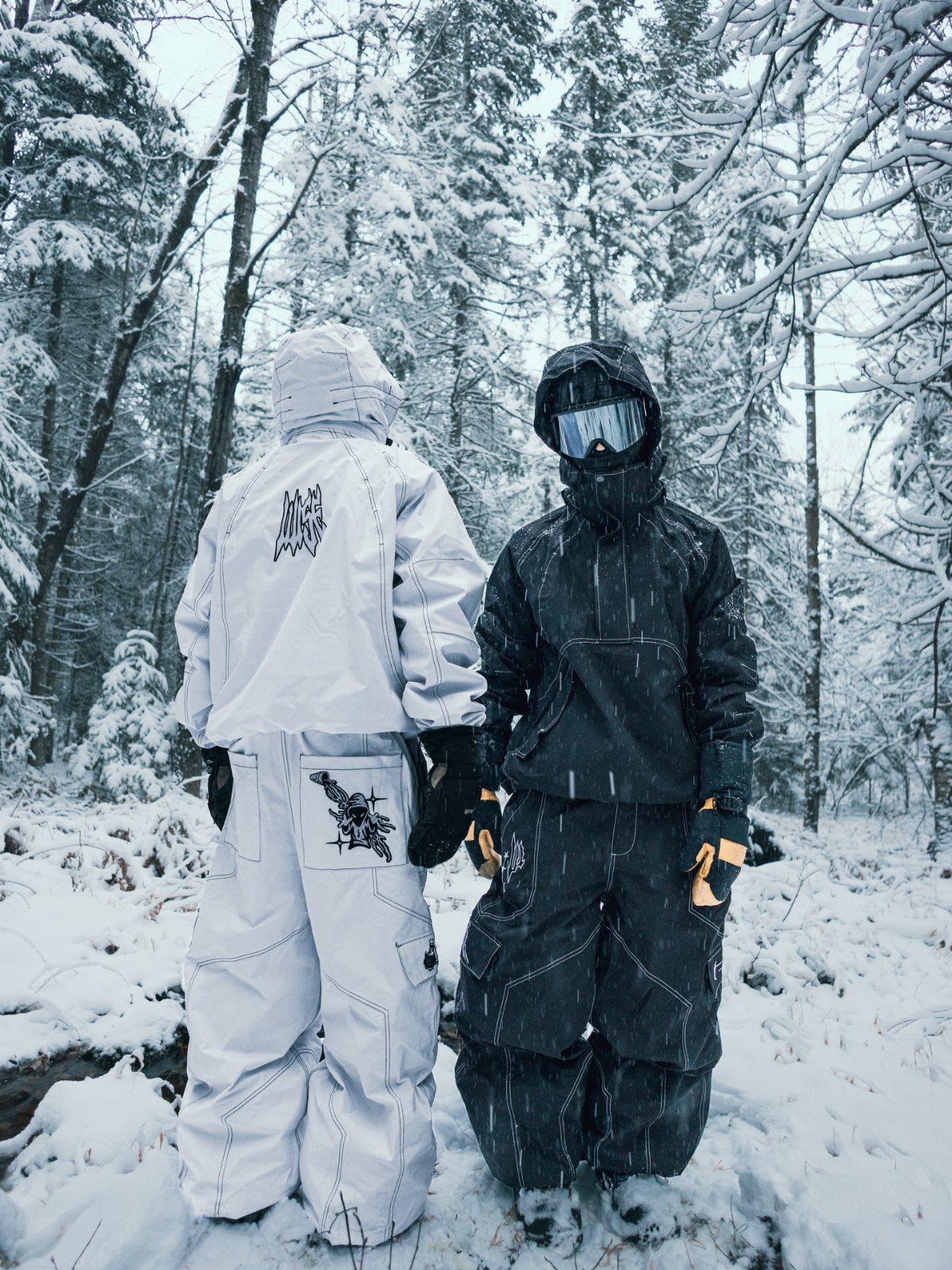 Two people wearing white and black snow gear standing in a snowy forest.