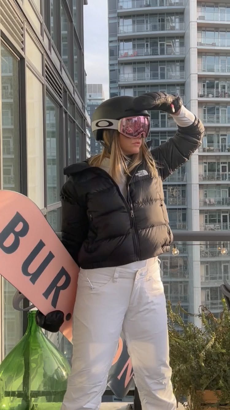 Woman in a black puffer jacket and white snow pants holding a pink snowboard.