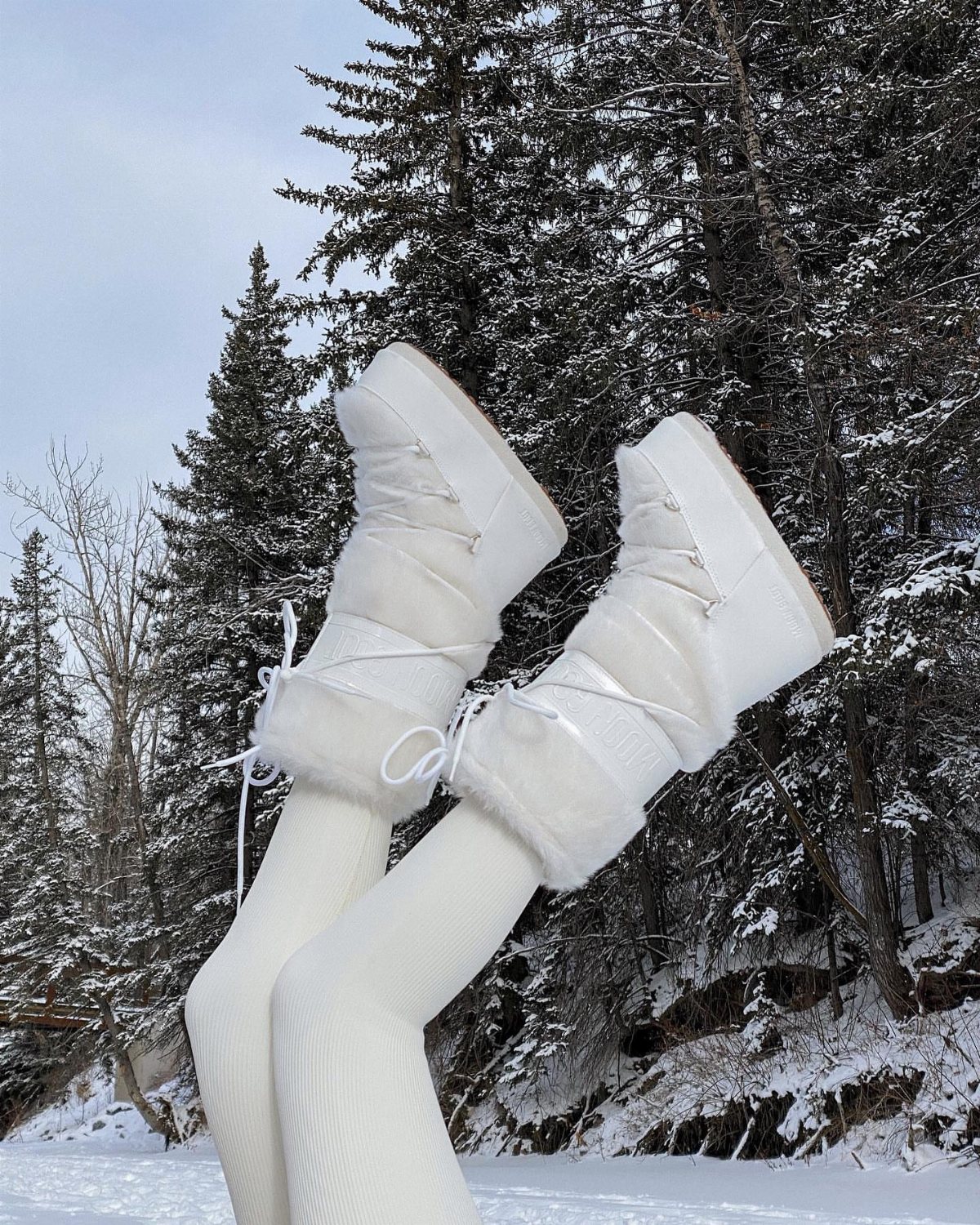 White fuzzy winter boots and white leggings raised against a snowy forest background.