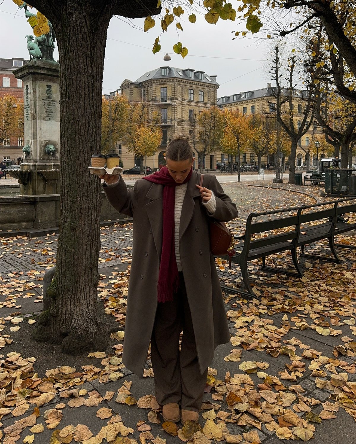 A woman in a long grey coat and burgundy scarf standing among fallen autumn leaves.