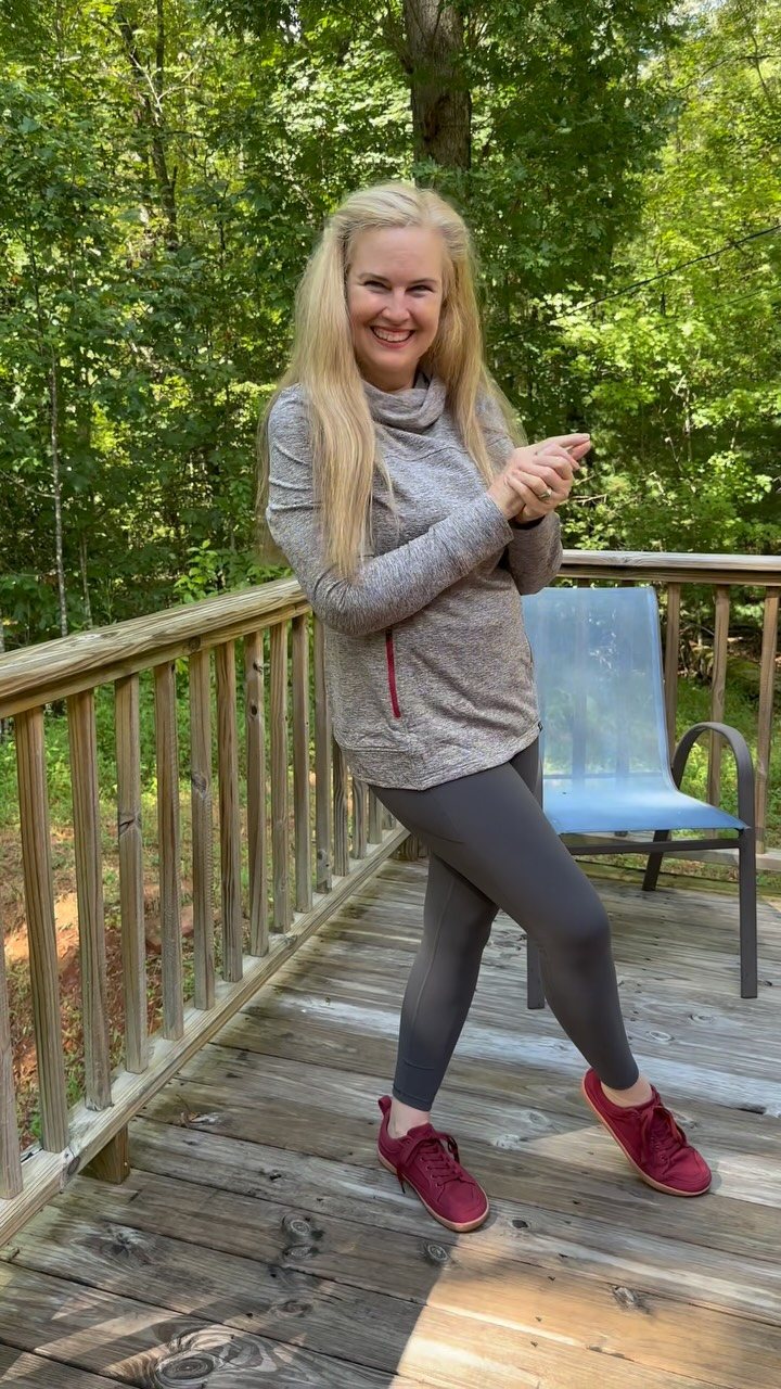Smiling blonde woman wearing a grey cowl neck pullover and dark leggings on a wooden deck.