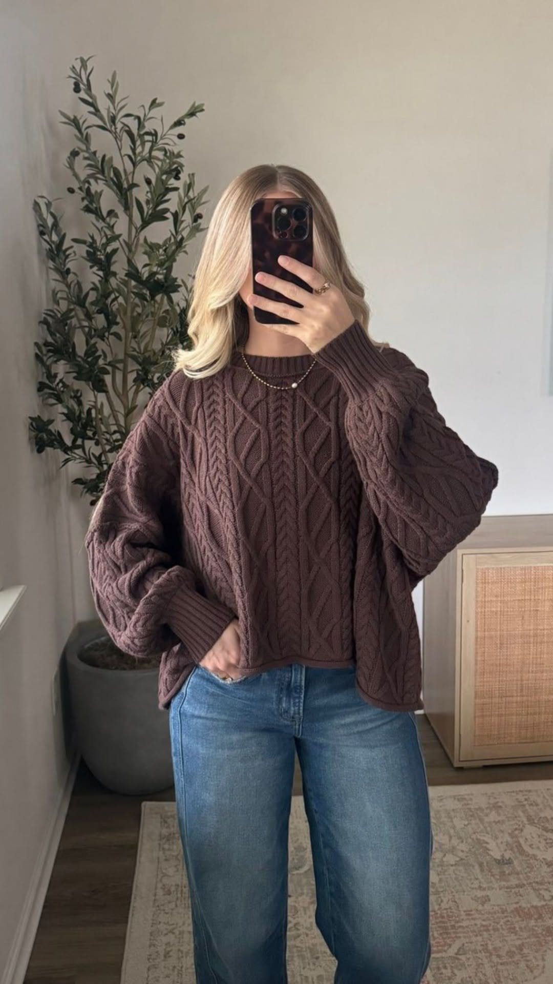 A woman posing in a mirror wearing a brown cable-knit chunky sweater and blue jeans.