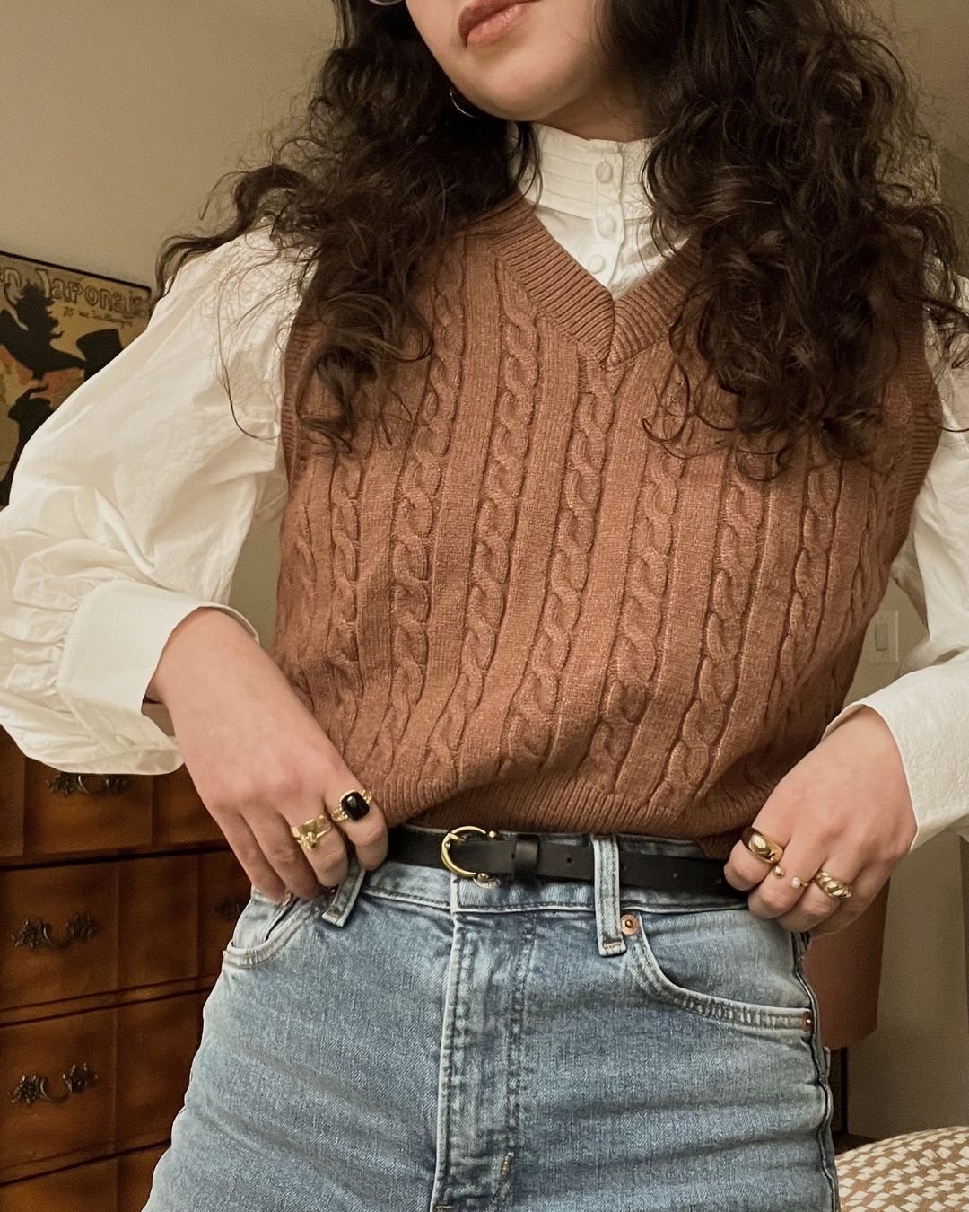 A person wearing a brown cable-knit sweater vest over a white blouse and blue jeans.