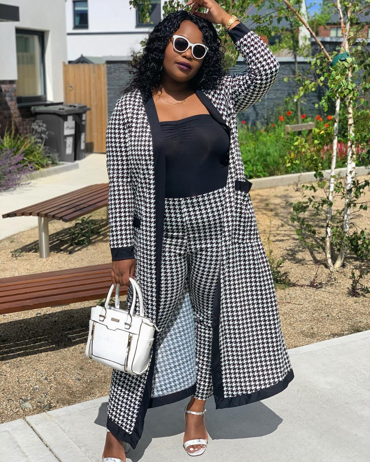 Woman in houndstooth duster coat, black top, trousers, white sunglasses, and carrying a white handbag.