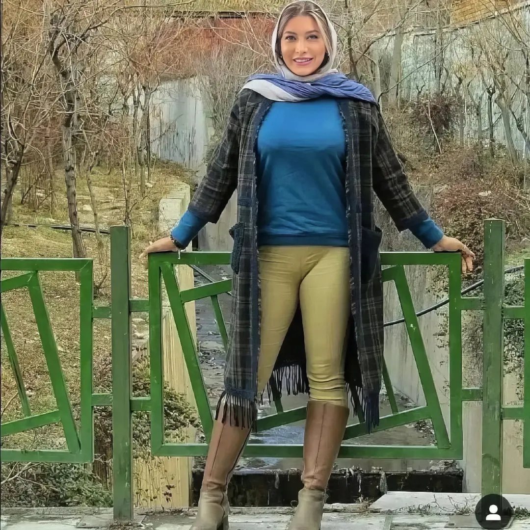 Woman in a plaid duster coat, blue shirt, tan pants, and tall tan boots standing by a fence.