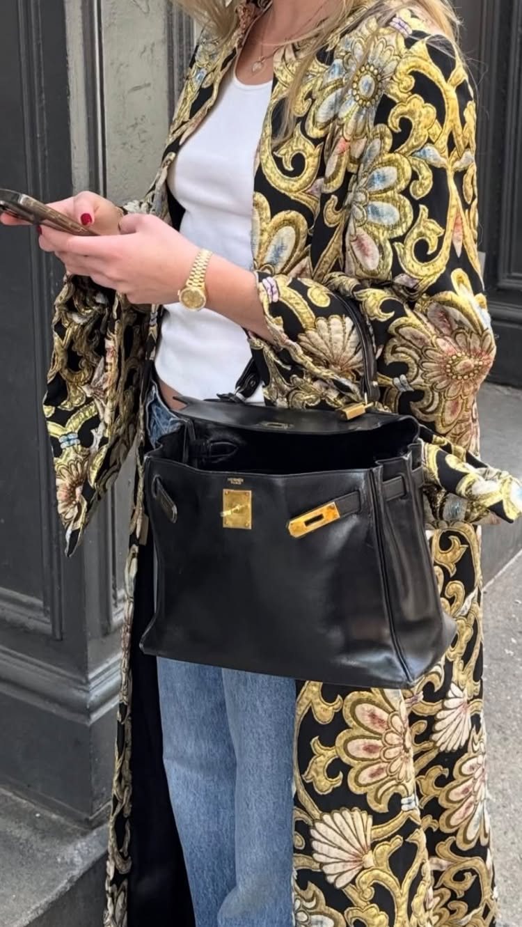 A person wearing a patterned gold and black duster coat with blue jeans and a black handbag.