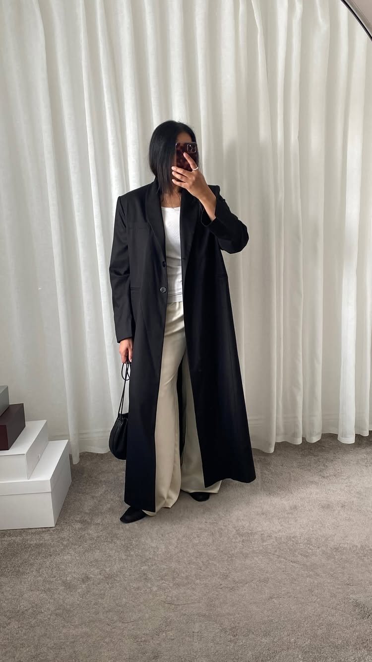 A person wearing a long black duster coat with cream wide-leg trousers standing before white curtains.