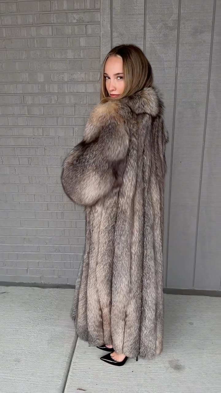 A woman wearing a long, voluminous grey fur coat and black heels standing against a grey wall.