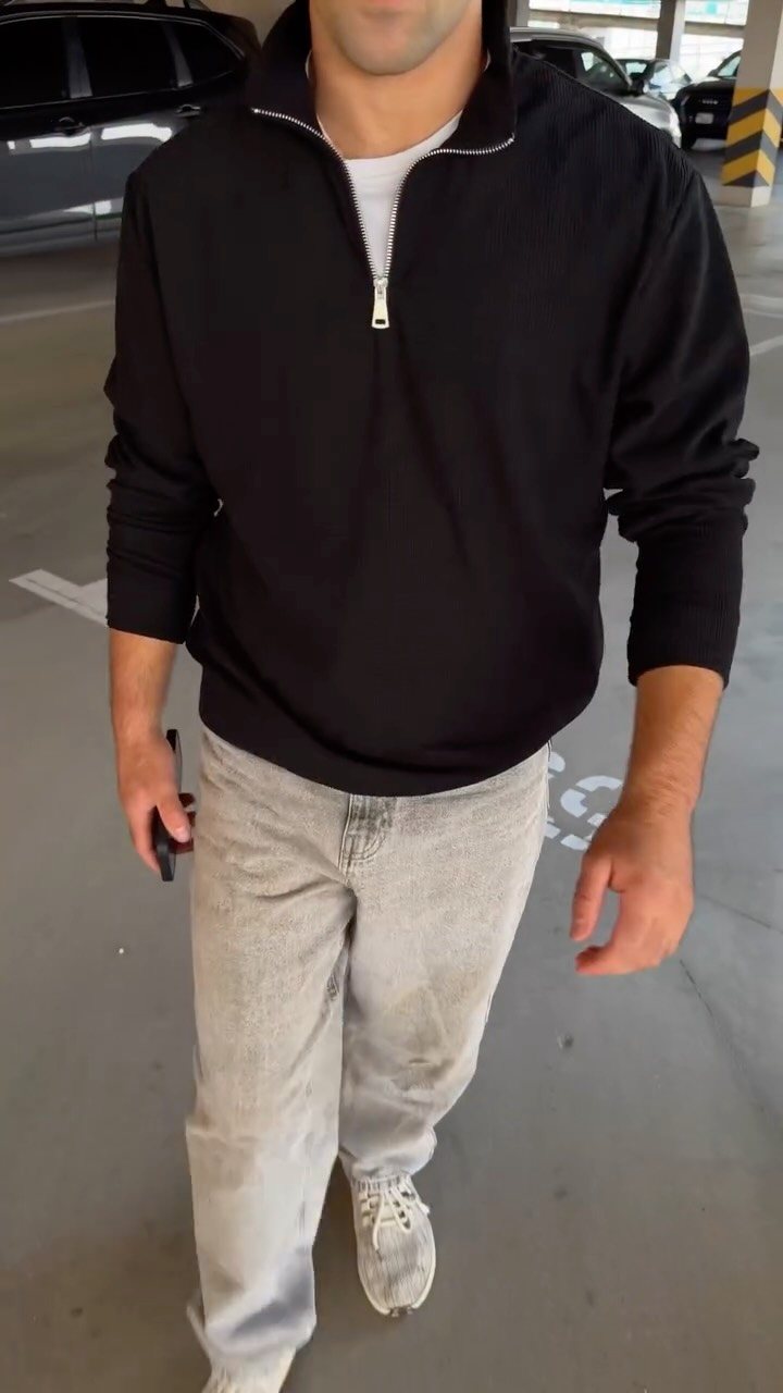 Man wearing a black quarter-zip sweatshirt and light grey denim jeans in a parking garage.