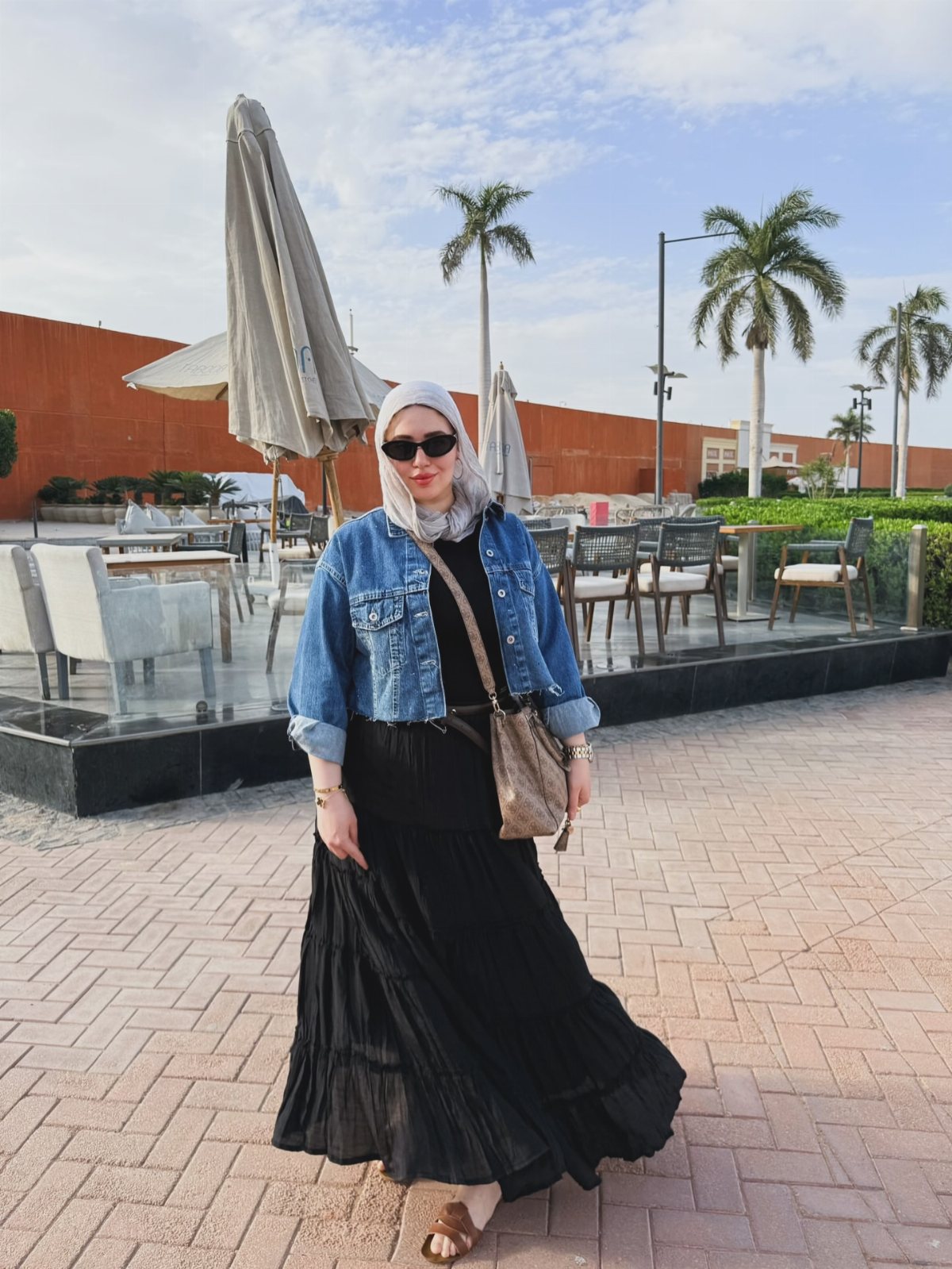 Woman in a blue denim jacket and long black dress standing on a brick patio.