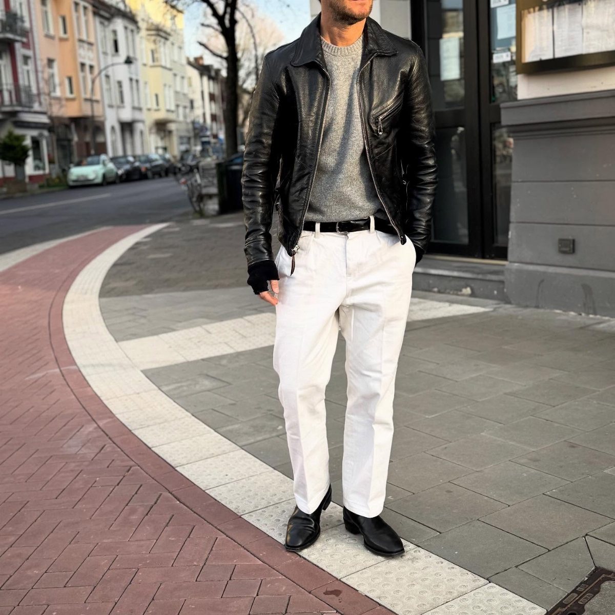 Man wearing a black leather jacket, grey sweater, and white trousers standing on a city street.
