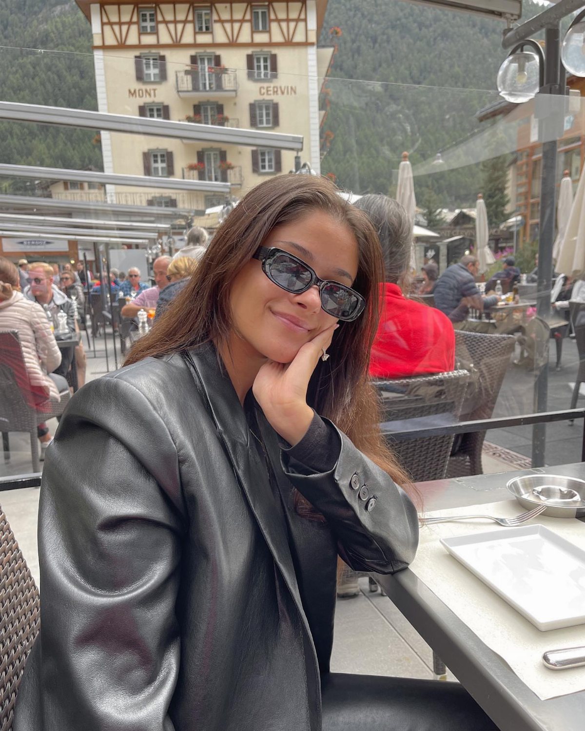 A smiling woman wearing sunglasses and a black leather blazer sitting at an outdoor cafe table.