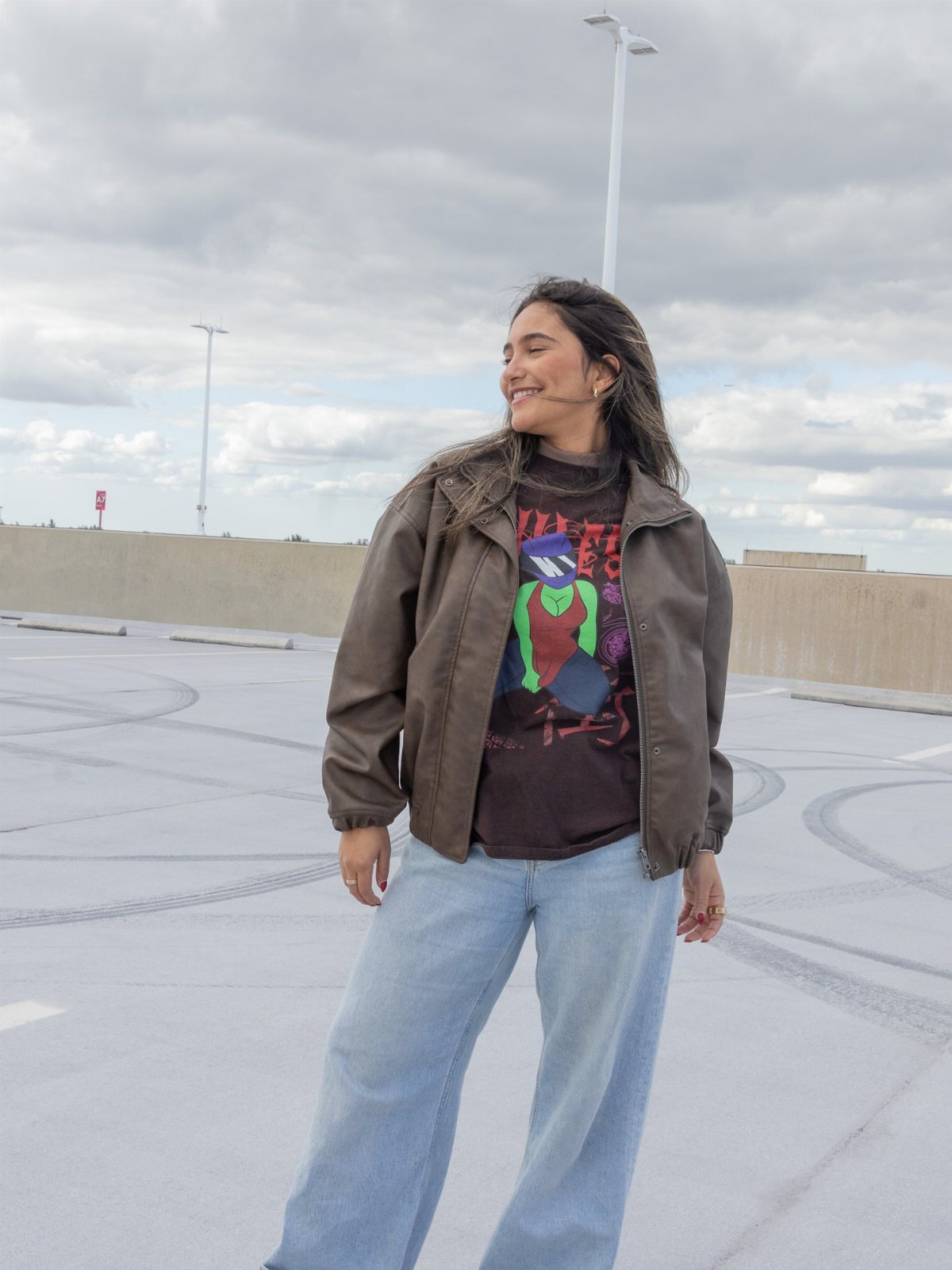 A woman wearing an olive bomber jacket, a graphic t-shirt, and light blue jeans.