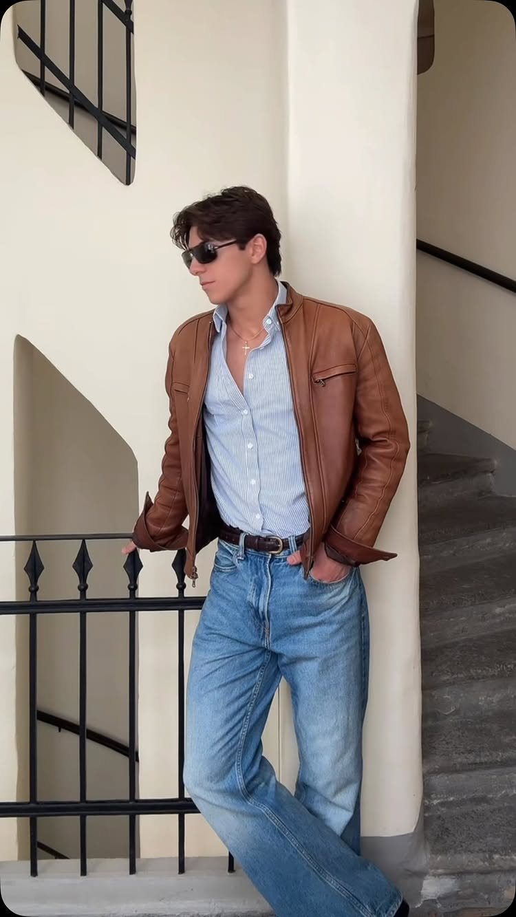 Man in a brown leather jacket and blue button-up shirt posing on a staircase.