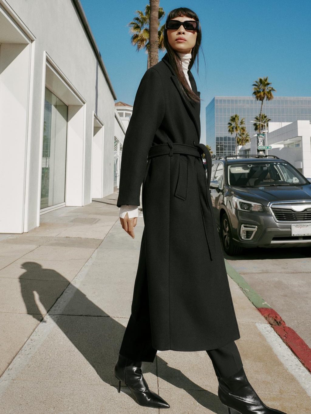 Woman in a long black belted coat and sunglasses walking on a sunny city sidewalk.