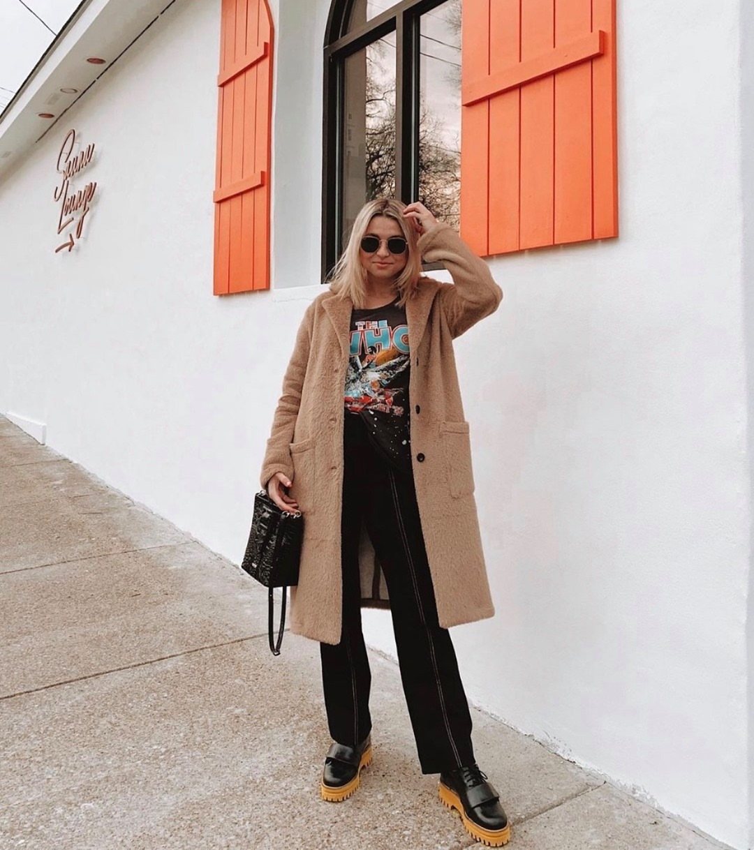 Woman wearing a long tan coat, graphic t-shirt, black pants, and chunky loafers.