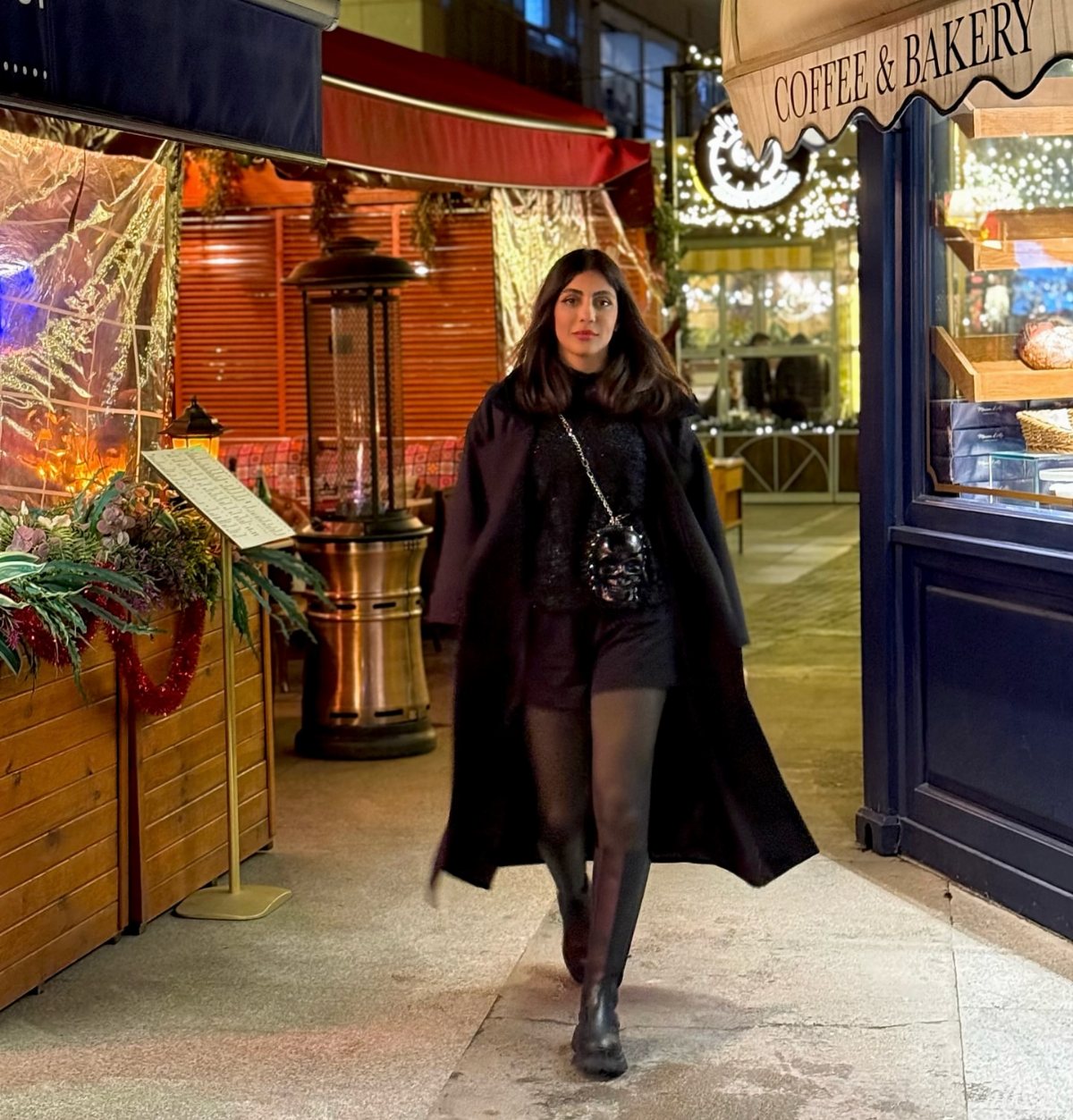 Woman in an all-black outfit with a long coat walking through a city street at night.