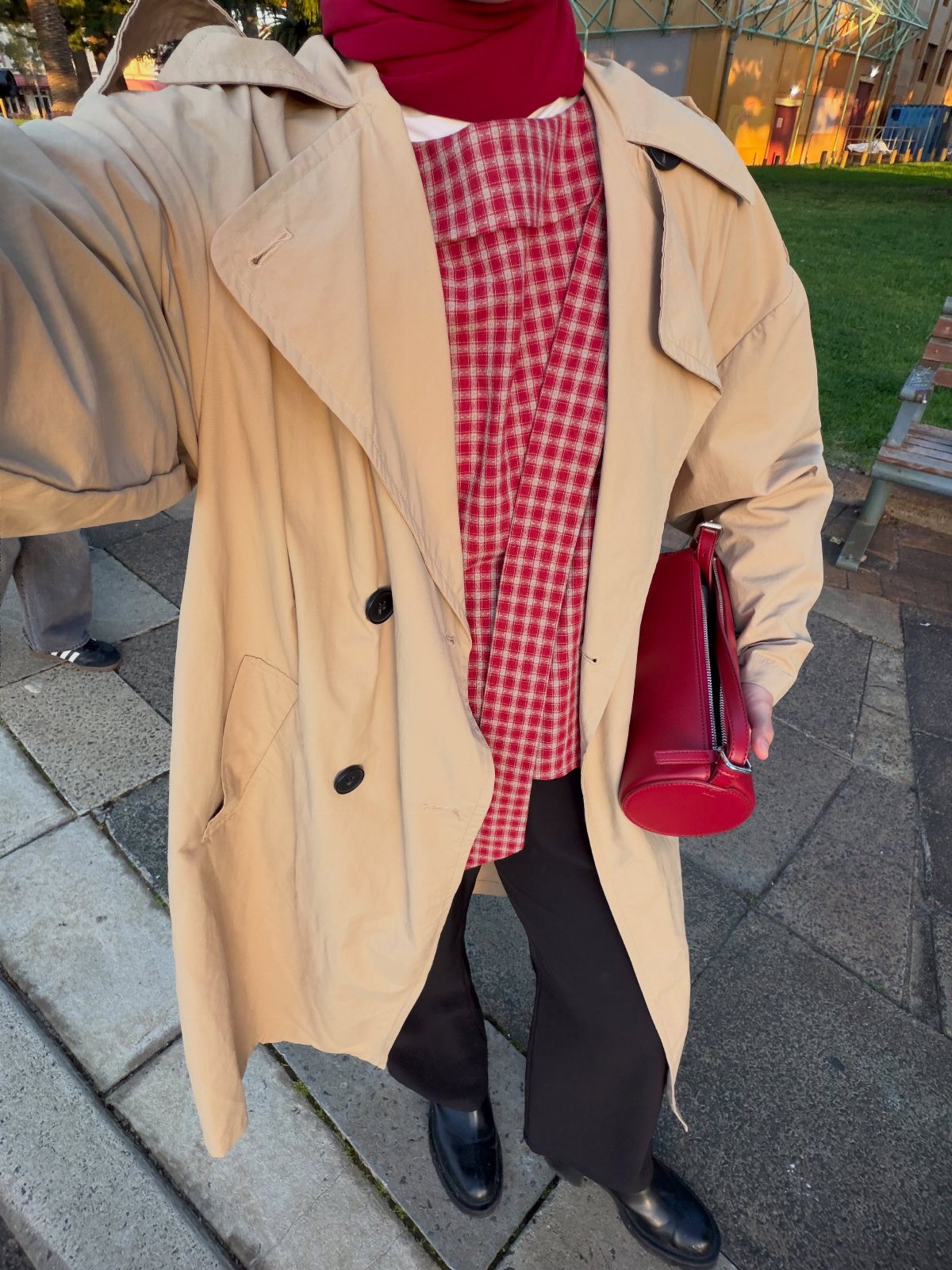 Person wearing a tan trench coat, red checkered scarf, and black trousers standing outdoors.