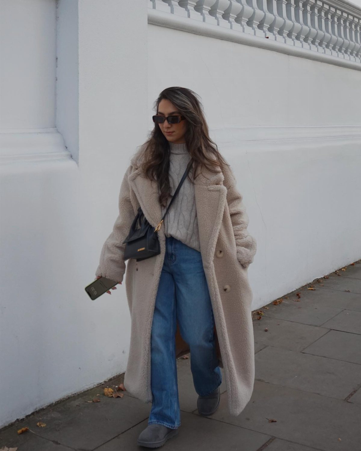 Woman wearing a long beige teddy coat, grey sweater, and blue jeans walking outside.