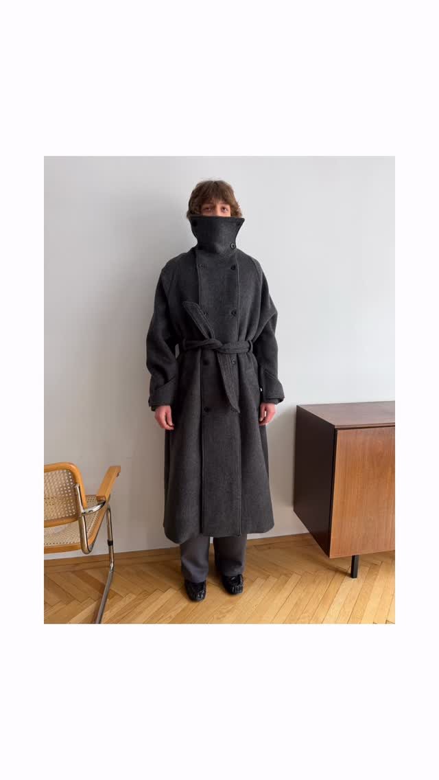 A person wearing an oversized charcoal grey wool coat with a belted waist in a minimalist room.