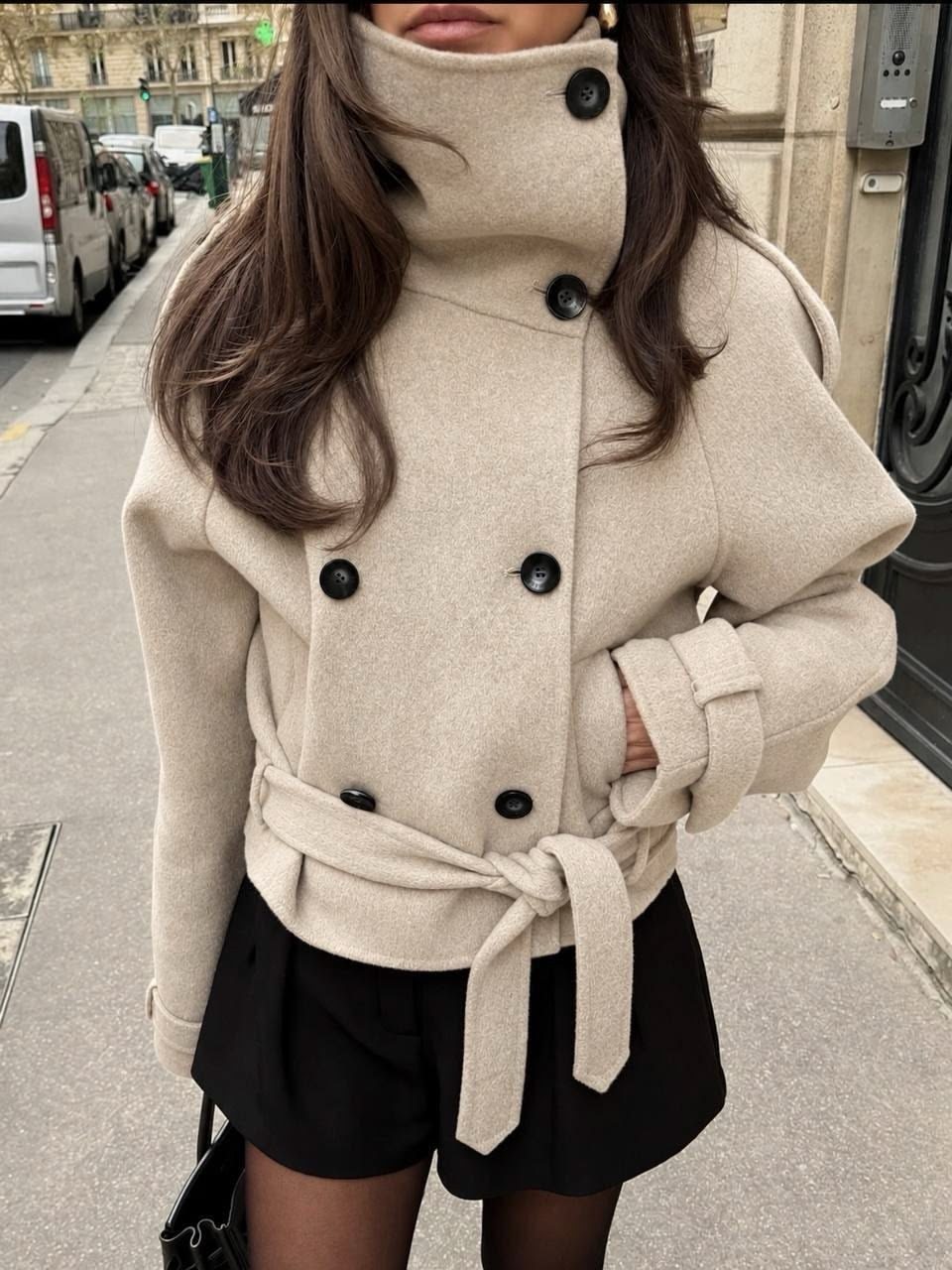 A woman wearing a thick off-white double-breasted wool coat with a tie belt on a city street.